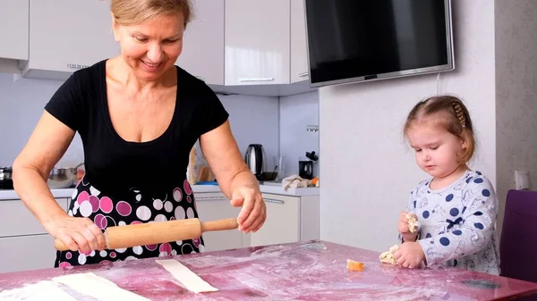 Grandma helps her daughter roll the dough in the kitchen to bake cookies. Grandma and daughter bake pizza together in the kitchen. The girl helps her mother roll out the dough with a rolling pin.