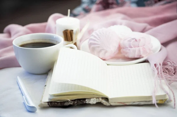 Handmade notebook and white pen. Coffee in the background, lush marshmallow close-up. Coffee mug in the background, candle tied with a ribbon with cinnamon and pink blurred background