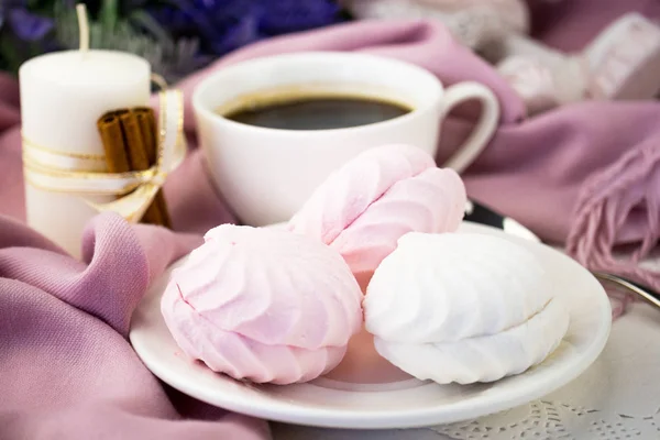 White and pink marshmallow on a saucer close-up. In the background is a pink scarf, a small cup of coffee and a white cinnamon-decorated candle