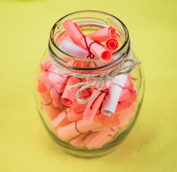 Round jar with notes. The notes are rolled up, pink and white. The jar is centered, the olive background is blurred