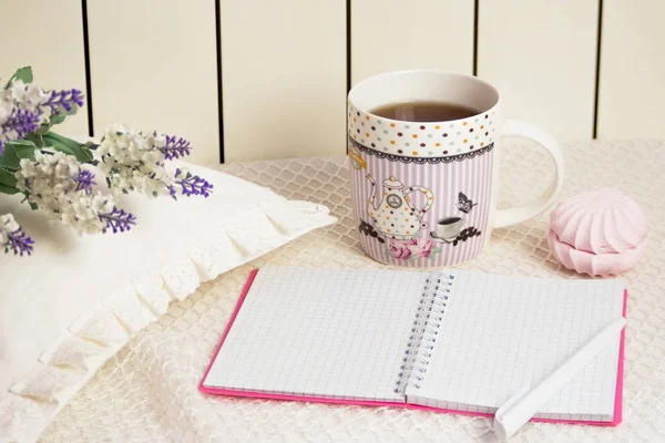 Notebook and pen on a white background. Near a mug of hot tea and sweets. Left cushion with lace and flowers
