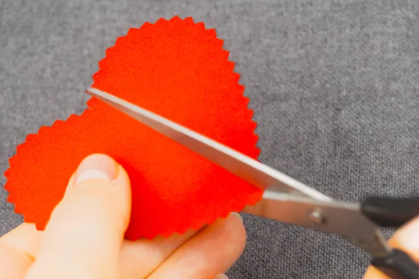 The hands of the girl cut the heart in half. Interesting backgrounds for Valentine\'s Day. Backgrounds with emotions, concept with a heart. Hands close up. Scissors cut a heart in half. Background blurred, gray fabric