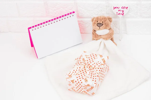 Toy bear on the background of a white brick wall. Gifts in a romantic package, next to a pink notebook with open pages, there is a place for signature. Vintage heart with the signature you are my love