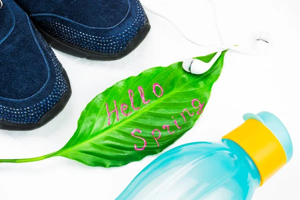 Doing fitness in the spring, sports concept. Green leaf with signature Hello Spring. Headset to listen to music. Blue, women\'s sneakers and sport gloves for the simulator. Eco bottle in bright colors for water. The background is light, large pane.
