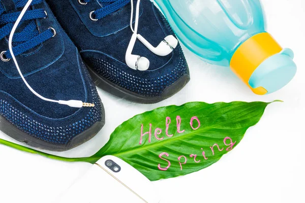 Doing fitness in the spring, sports concept. Green leaf with signature Hello Spring. Headset to listen to music. Blue, women\'s sneakers and sport gloves for the simulator. Eco bottle in bright colors for water. The background is light, large pane.