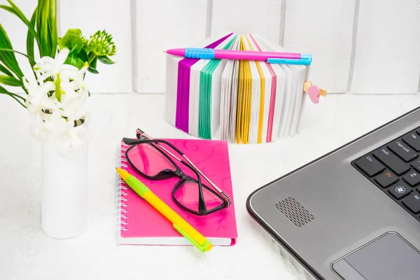 The working environment in the women\'s office. A set of stationery, a notebook business woman, multi-colored pens for writing, a pack of stickers, glasses and a laptop computer on a light background with a composition of white flowers. Concept female