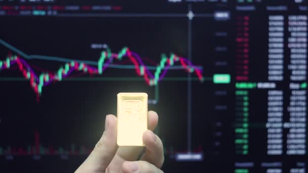 Eine Hand Hält Einen Goldbarren Vor Dem Hintergrund Eines Handelspreisdiagramms — Stockvideo