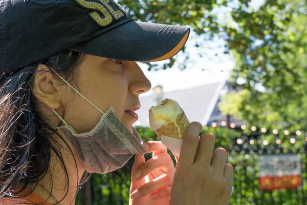 A young girl of Caucasian appearance eats an ice cream cone in a medical mask on the street during a quarantine for a coronavirus infection in summer. The weakening of the quarantine in the countries