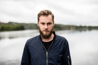 Mavi bir ceket ve kot, woods çevreleyen ile büyük bir göl ile ayakta dışında giyen adam dalgın bir sakallı.