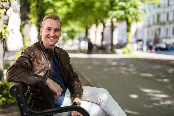 Relaxed young man sitting down on a bench outdoors wearing a brown leather jacket looking at camera smiling. Sunny summer day.