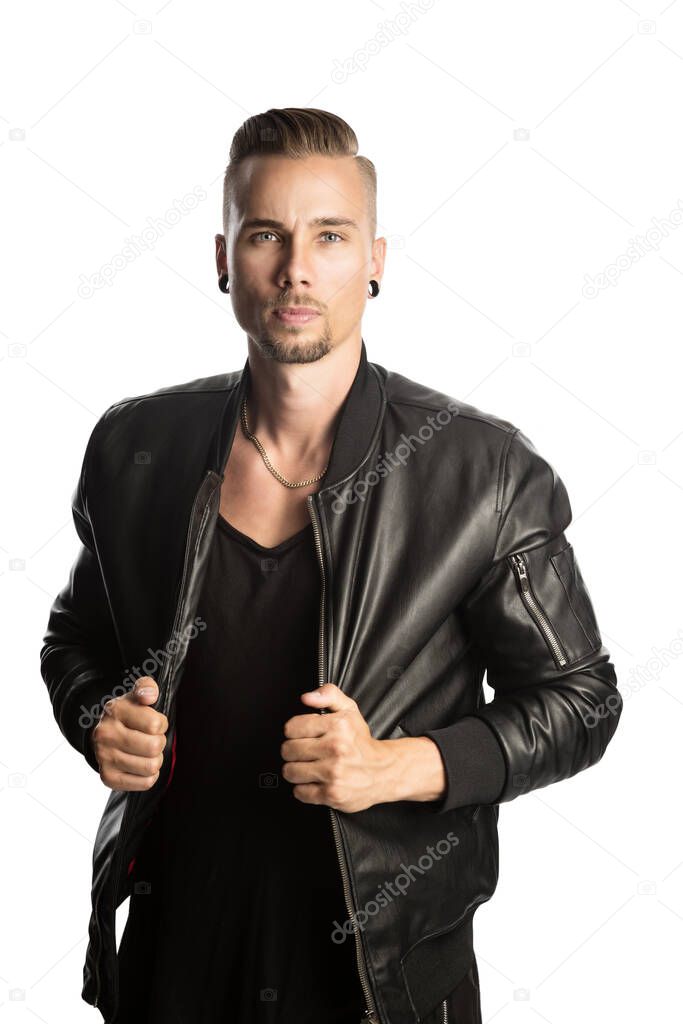 Attractive young beardedman wearing a trendy black leather jacket and black shirtlooking angry at the camera with both hands holdinghis jacket and standing against white background.