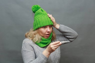 Gri kazak endişeli genç kadın, atkı alnına el koymak, gri arka plan üzerinde izole termometre tutun.