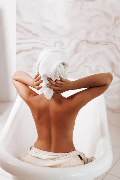 Body care concept. Back view of young adult woman taking bath in comfortable bathroom.