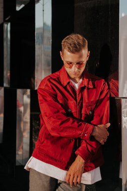 Gündüz vakti modern binanın cam duvarına dayanan güneş gözlüklü, modaya uygun giyinmiş genç bir erkek. Büyük bir şehrin caddesinde şık giyinmiş güzel bir adam.