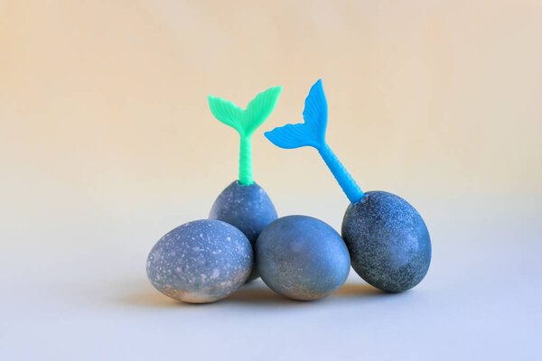 Четыре голубых пасхальных яйца лежат на желтом фоне. Синие и зеленые хвосты русалок торчат из яиц
.