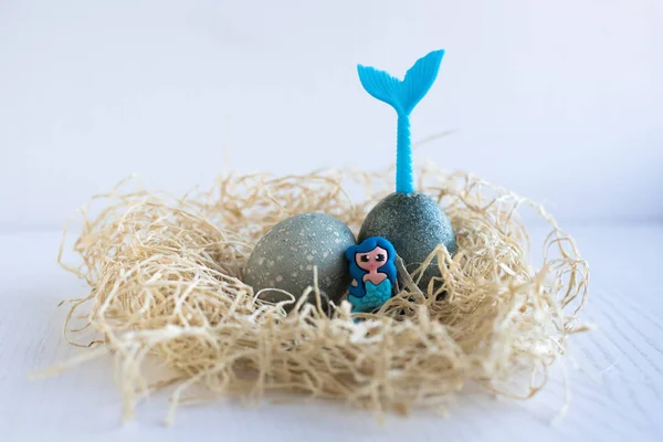 Easter eggs lie in a nest on a white wooden background. Tail of mermaid stick out of egg. Mermaid sitting in the nest