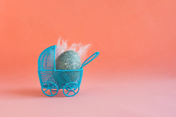 Голубое пасхальное яйцо в голубой коляске на красном коралловом фоне
