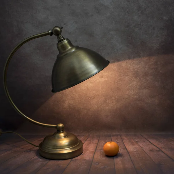 A table lamp illuminates a tangerin laying on a wooden table, dark background