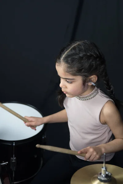 A 5 year old girl of British Indian ethnicity - practicing on the drums and reading music notes - on a dark black background.