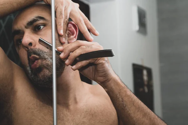 A bald, British Asian man, grooming and sculpting his beard at home in the bathroom, in front of the mirror. He is using a traditional cut throat razor or a barber\'s razor. This can be used to theme issues around maleness, dating and middle age.