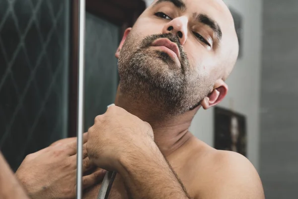 A bald, British Asian man, grooming and sculpting his beard at home in the bathroom, in front of the mirror. He is using a traditional cut throat razor or a barber\'s razor. This can be used to theme issues around maleness, dating and middle age.