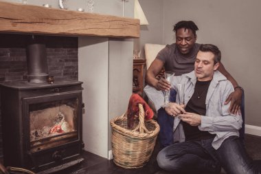 A multi ethnic gay couple sits at home by the fireplace - in Britain - spending a cozy time together.  clipart