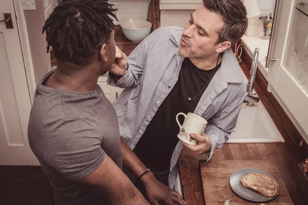 A gay couple sharing stories from their day at work over a slice of toast and a cup of tea and coffee in the evening.