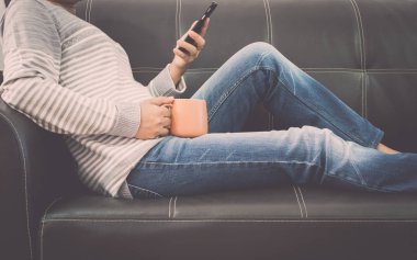 Cep telefonu kullaranarak ve bir fincan kahve evde tutan deri koltukta kadındır.