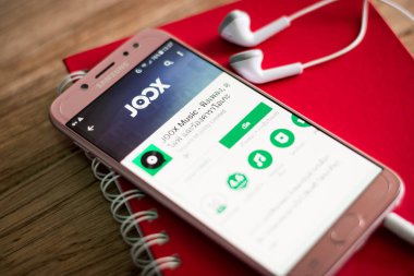 Phuket, Tayland - 23 Ağustos 2018: Ekrandaki Joox müzik uygulamasıyla ahşap masa üstünde earsphone ve kırmızı defter Samsung Galaxy J7 pro smartphone başlattı.