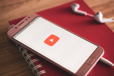 Phuket, Tayland - 23 Ağustos 2018: Akıllı telefon Samsung Galaxy J7 pro açýk apps youtube uygulama earphones ve ahşap masa üstünde defter ile ekranda.