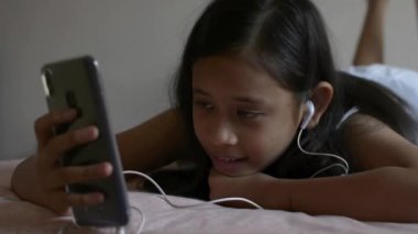 Yatakta yatan kulaklıkile cep telefonu kullanarak rahat elbiseli sevimli kız.