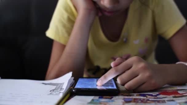 机の上で宿題をしながら情報を見つけるために携帯電話を使用して若い女の子 技術と教育の概念 — ストック動画