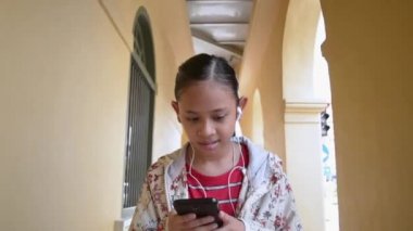 Chino portekiztarzı bina koridorunda yürürken kulaklıkile cep telefonundan müzik dinlesevimli kız.