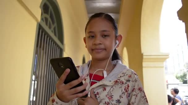 チノポルトガル風の建物の廊下を歩きながらイヤホンで携帯電話から音楽を聴くかわいい女の子 — ストック動画