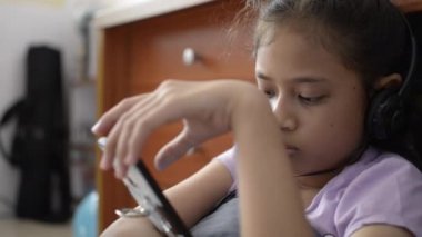 Küçük asyalı kız kulaklık ile cep telefonu kullanarak ve evde video izlerken.