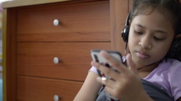 小亚洲女孩使用手机与耳机和观看视频在家里 — 图库视频影像