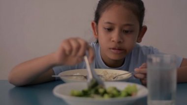 Asyalı tatlı kız akarsu pirinçle öğle yemeği yiyor ve masada kızarmış brokoli pişiriyor, sağlıklı yiyeceklerle yemek yiyor..