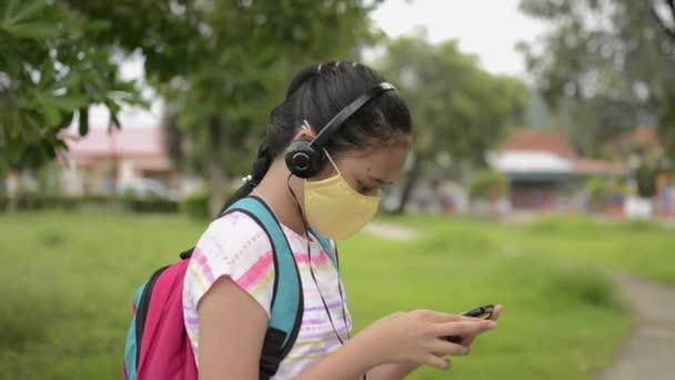 ヘッドフォンを着用し 公園に立って保護顔マスクのアジアの小学生 女性のティーンエイジャーは 住宅街で携帯電話を使用してバックパックで コロナウイルス対策 回転ショット — ストック動画