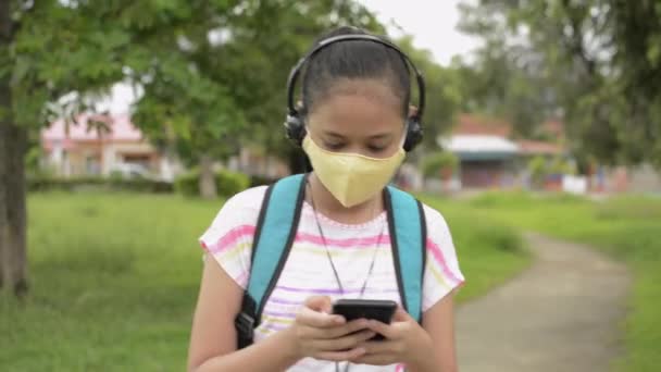 ヘッドフォンを着用し 住宅街の公園で歩く保護顔マスクのアジアの中等学校の女の子 帰宅途中 携帯電話をバックパックにした女子10代 コロナウイルス対策 — ストック動画
