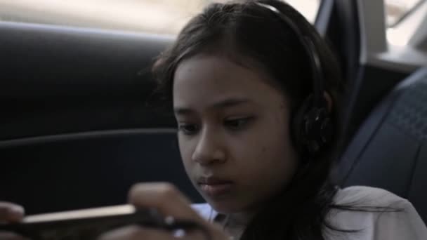 可爱的亚洲女孩在开车旅行时戴着耳机和使用手机 坐在汽车后座上观看网上电影的女青少年 路途中的娱乐 — 图库视频影像