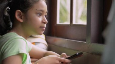 Pencerenin kenarında oturan ve cep telefonu kullanan sıradan giysili tatlı Asyalı kız. Genç kız sosyal medya için akıllı telefondan mesaj atıyor. İletişim için kablosuz teknoloji.