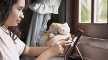 Pencerenin kenarında oturup dijital tablet kullanan sıradan giysili tatlı Asyalı kız. Dişi ergen dijital cihazda eğitici bilgiler arıyor. İletişim için kablosuz teknoloji.