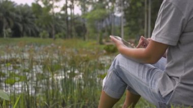 Normal giysili Asyalı kadın doğal göletin yanındaki çimlerin üzerinde oturuyor ve sosyal güvenlik için cep telefonunu kullanıyor. Döndürme atışı. Kablosuz teknoloji. Sosyal iletişim.