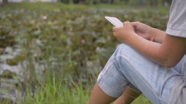 Femme asiatique en robe décontractée est assis sur l'herbe près de l'étang naturel et textos sur téléphone mobile pour social en ligne. Technologie sans fil. Communication sociale.