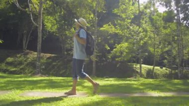 Hasır şapka ve sırt çantası takan Asyalı kadın turist, sabah güneş ışığı altında orman parkında çimlerin üzerinde rahatça yürüyor. Yan görüş takip çekimi. Ağır çekim. Namtok Raman Orman Parkı. Phang Nga Eyaleti. Tayland.
