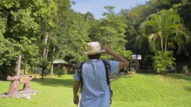 Hasır şapka ve sırt çantası takan Asyalı kadın turist, sabah güneş ışığı altında orman parkında çimlerin üzerinde rahatça yürüyor. Arka görüş takip çekimi. Ağır çekim. Namtok Raman Orman Parkı. Phang Nga Eyaleti. Tayland.