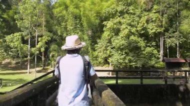 Hasır şapkalı ve sırt çantalı Asyalı kadın turist eski beton köprüde yürüyor ve cep telefonuyla fotoğraf çekiyor. Gezginler yaz boyunca orman parkında gün ışığında vakit geçirmekten zevk alırlar. Phang Nga Eyaleti. Tayland.