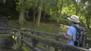 Hasır şapkalı ve sırt çantalı Asyalı kadın turist eski beton köprüde yürüyor ve cep telefonuyla fotoğraf çekiyor. Gezginler yaz boyunca orman parkında gün ışığında vakit geçirmekten zevk alırlar. Phang Nga Eyaleti. Tayland.