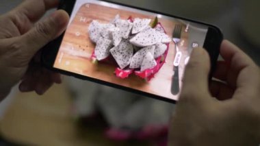 Kadının elleri, ahşap kesme tahtasının üzerinde cep telefonu kullanarak hazır ejderha meyvelerinin fotoğrafını çekiyor. Yemek fotoğrafçılığı. Sağlıklı beslenme. Taylandlı kamera uygulaması.