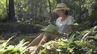 Asyalı kadın hasır şapka takıyor ve bir ağacın altında deftere yazı yazıyor. Biyolog, tropikal ormanlardaki doğal verileri ve ekosistemi not alıyor. Doğa ve koruma. Döndürme çekimi.